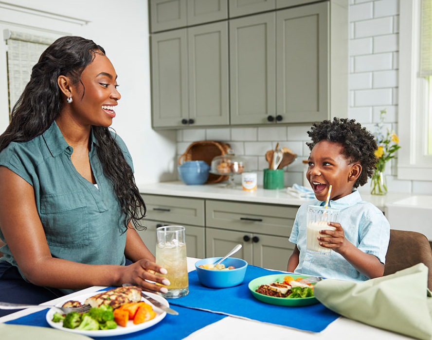Young boy eating at the table in a bright kitchen, smiling and holding a glass of EleCare Jr. Mom is smiling back at him.