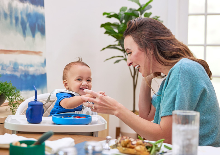 Mom smiling and holding her baby’s hand. The baby is sitting in a high chair with food and a sippy cup of EleCare.
