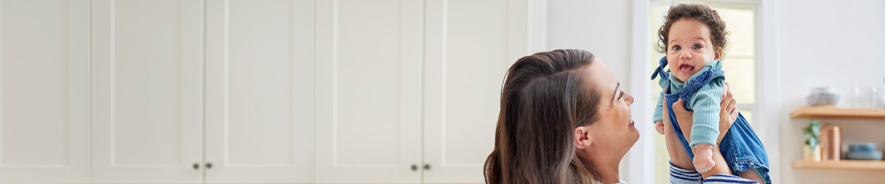 Woman standing in kitchen and smiling while holding up happy, smiling baby wearing blue.