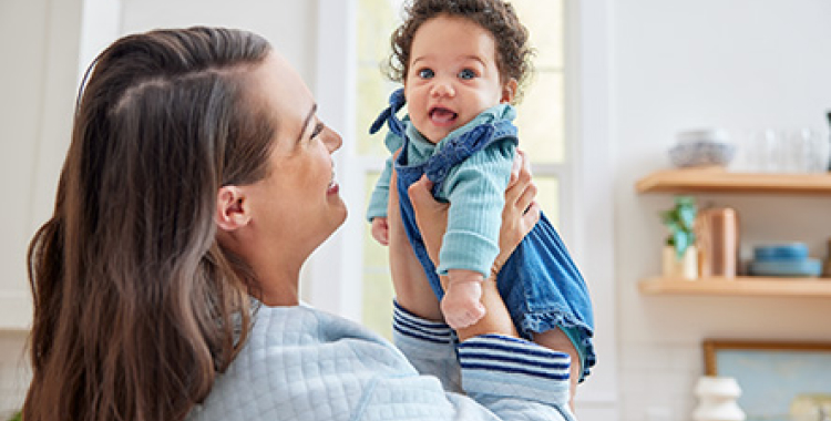 Woman standing in kitchen and smiling while holding up happy, smiling baby wearing blue.