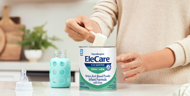Close view of a woman in beige, in the kitchen, scooping EleCare Infant Formula from the can into a baby bottle with water.