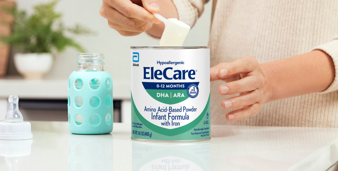 Close view of a woman in beige, in the kitchen, scooping EleCare Infant Formula from the can into a baby bottle with water.
