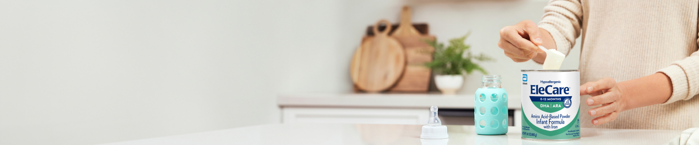 Close view of a woman in beige, in the kitchen, scooping EleCare Infant Formula from the can into a baby bottle with water.