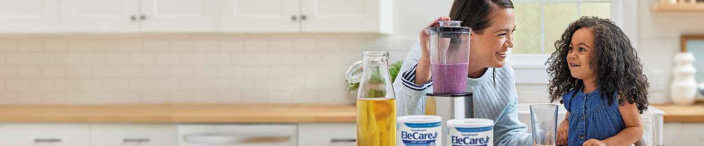 Woman in kitchen making a purple smoothie in a blender using EleCare Jr. Young girl watching and waiting for the smoothie.