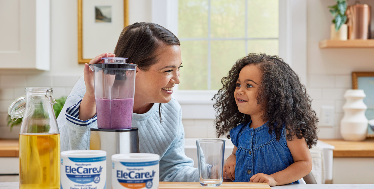 Woman in kitchen making a purple smoothie in a blender using EleCare Jr. Young girl watching and waiting for the smoothie.