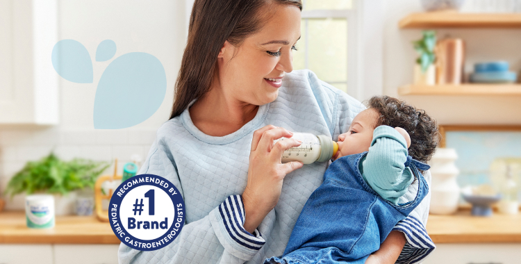Mom standing in kitchen, holding her baby daughter, who is dressed in blue and being fed a bottle of EleCare.