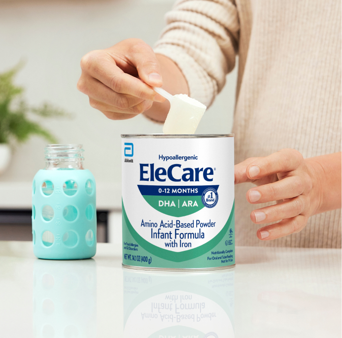 Close view of a woman in beige, in the kitchen, scooping EleCare Infant Formula from the can into a baby bottle with water.