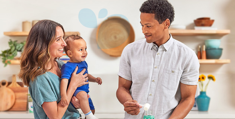 Mom in kitchen holding a baby boy in blue, while Dad is making a bottle of EleCare.