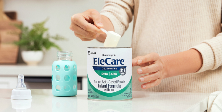 Close view of a woman in beige, in the kitchen, scooping EleCare Infant Formula from the can into a baby bottle with water.