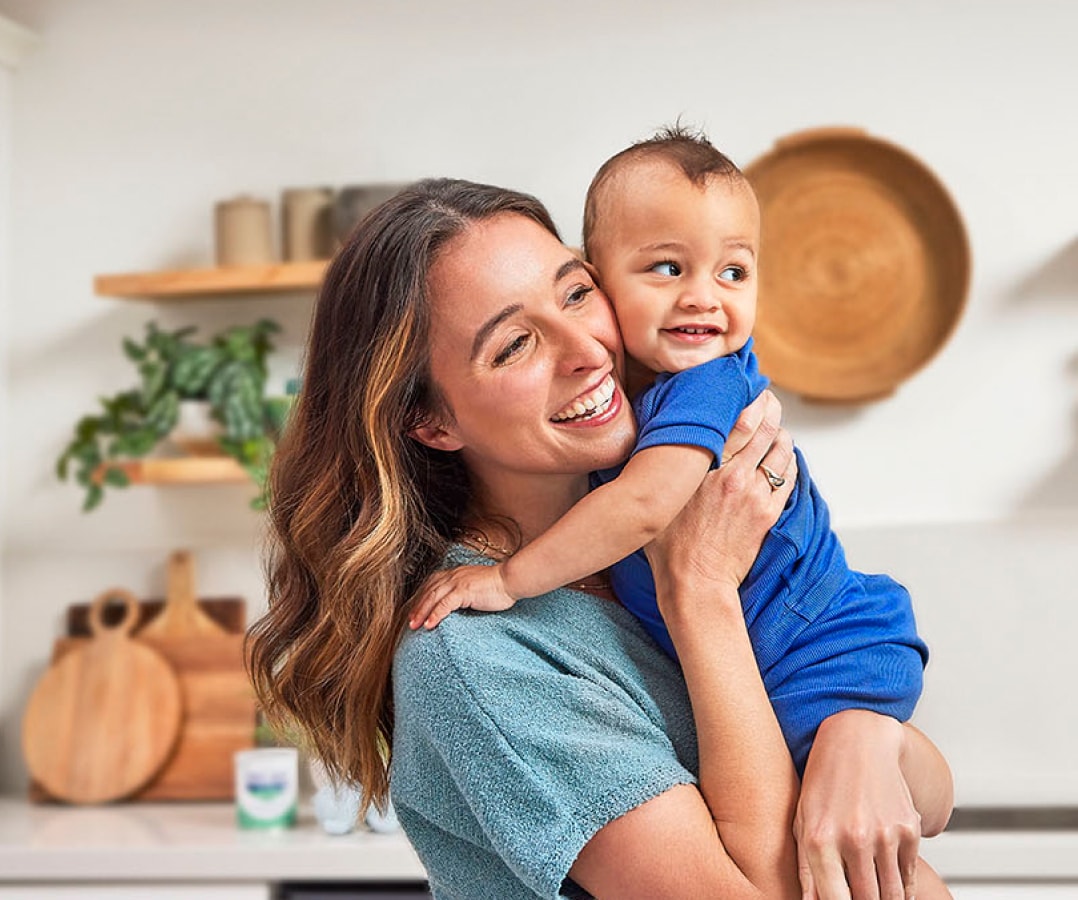 Woman holding her baby son, cheek to cheek, in the kitchen. Both are dressed in tones of blue and smiling.