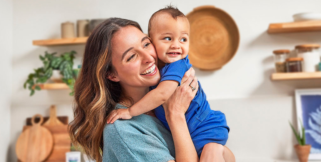 Woman holding her baby son, cheek to cheek, in the kitchen. Both are dressed in tones of blue and smiling.