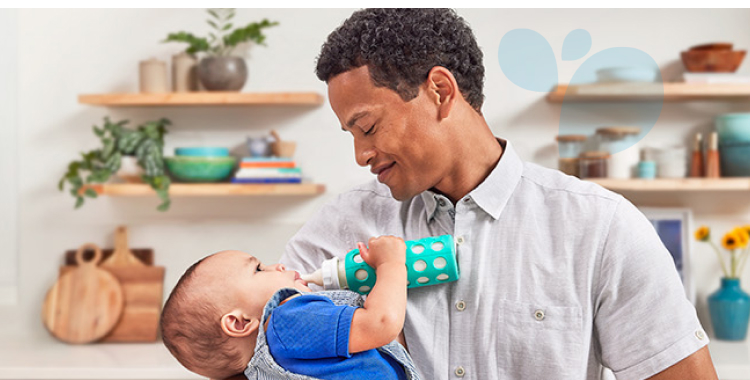 Man standing in kitchen and holding his baby boy, who is dressed in blue and drinking a bottle of EleCare.