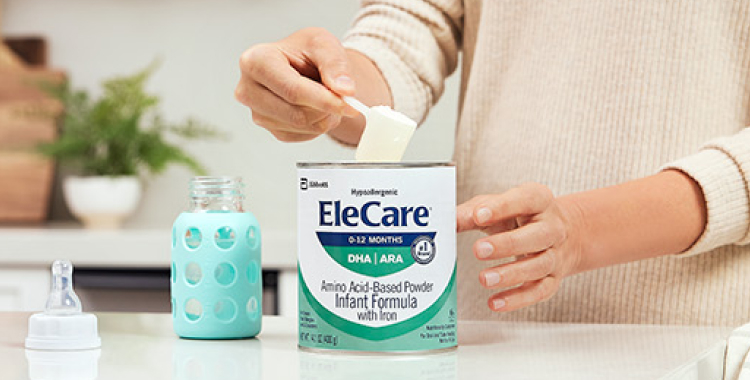 Close view of a woman in beige, in the kitchen, scooping EleCare Infant Formula from the can into a baby bottle with water.