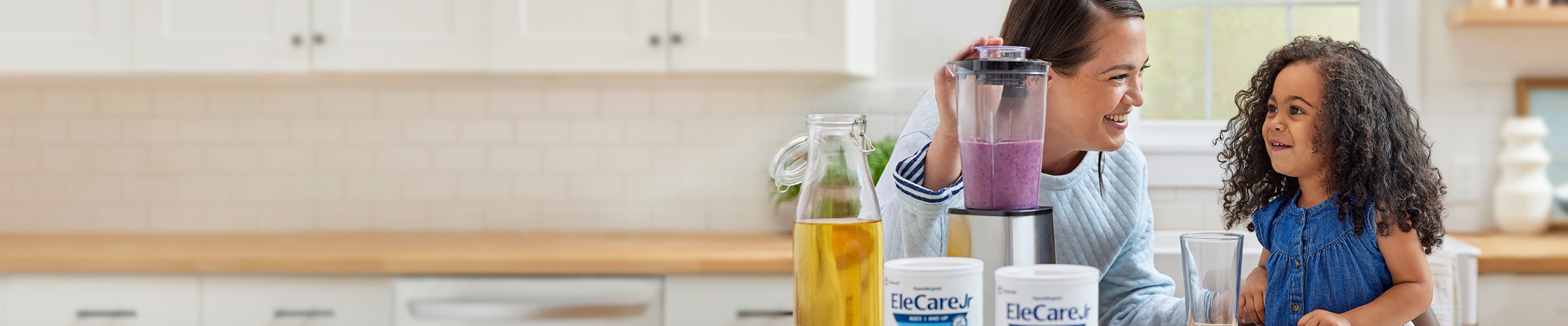 Woman in kitchen making a purple smoothie in a blender using EleCare Jr. Young girl watching and waiting for the smoothie.