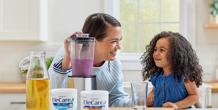 Woman in kitchen making a purple smoothie in a blender using EleCare Jr. Young girl watching and waiting for the smoothie.
