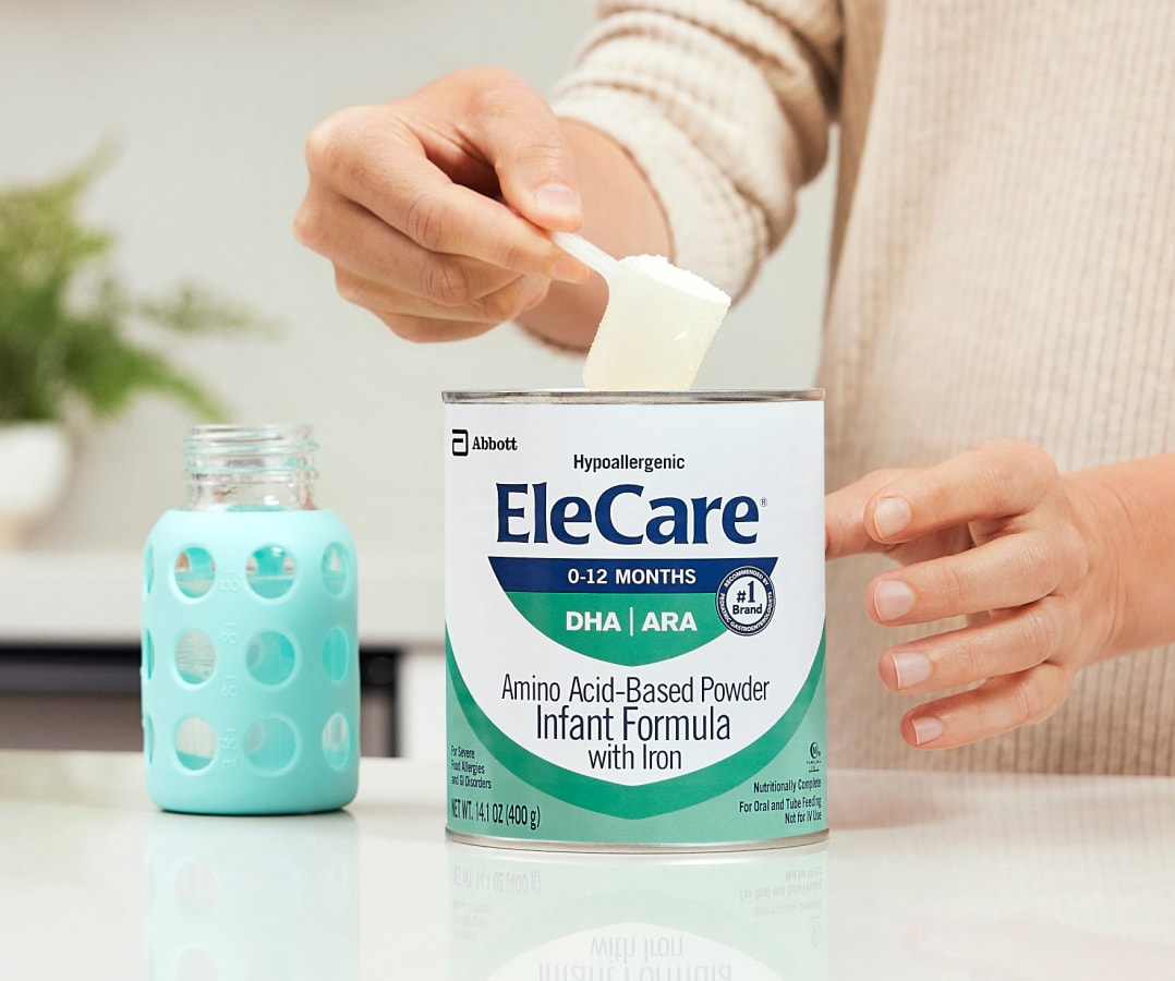 Close view of a woman in beige, in the kitchen, scooping EleCare Infant Formula from the can into a baby bottle with water.