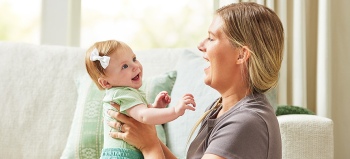 Woman in front of couch in living room, holding baby girl up; both are smiling at each other.