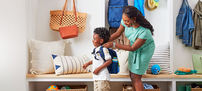 Woman in bright mudroom of house, placing blue backpack on young boy; both are smiling.