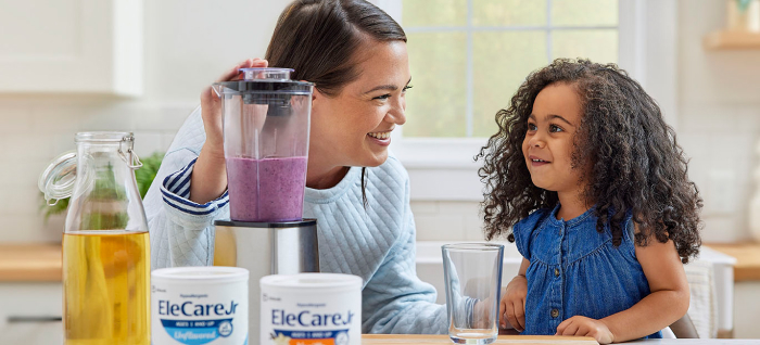 Woman in kitchen making a purple smoothie in a blender using EleCare Jr. Young girl watching and waiting for the smoothie.