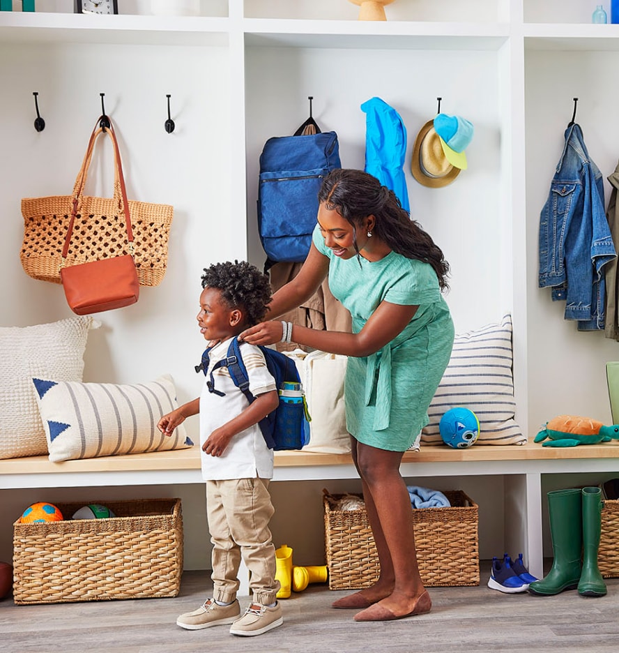 Woman in bright mudroom of house, placing blue backpack on young boy; both are smiling.