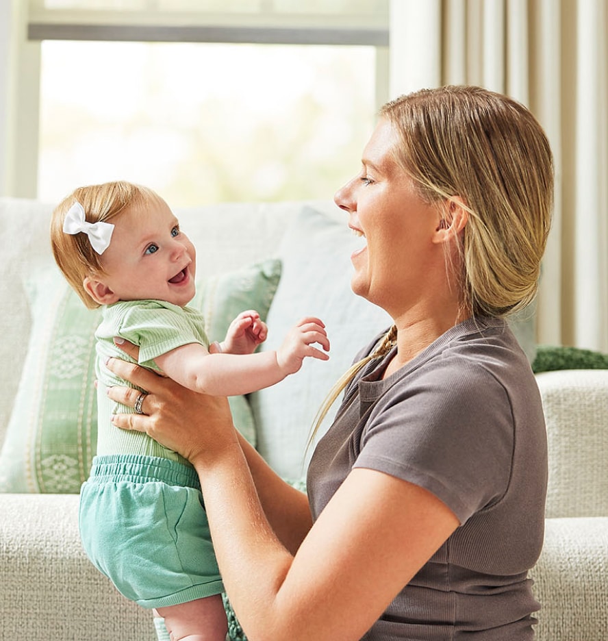Woman in front of couch in living room, holding baby girl up; both are smiling at each other.