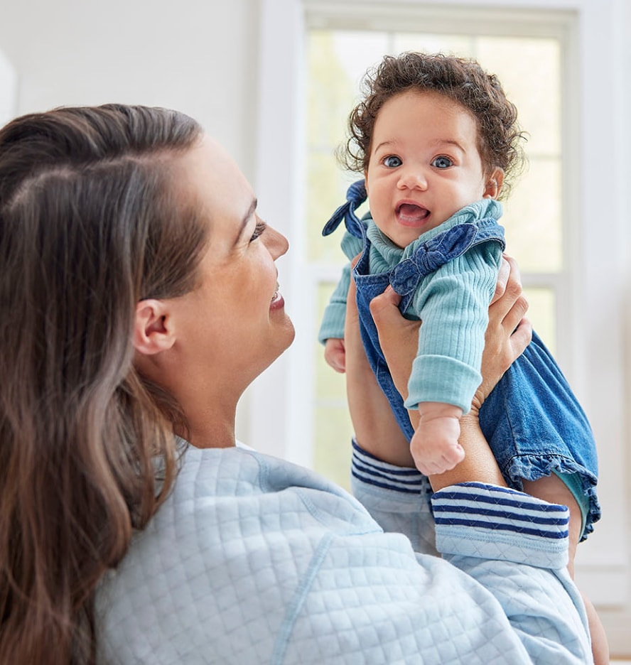 Woman standing in kitchen and smiling while holding up happy, smiling baby wearing blue