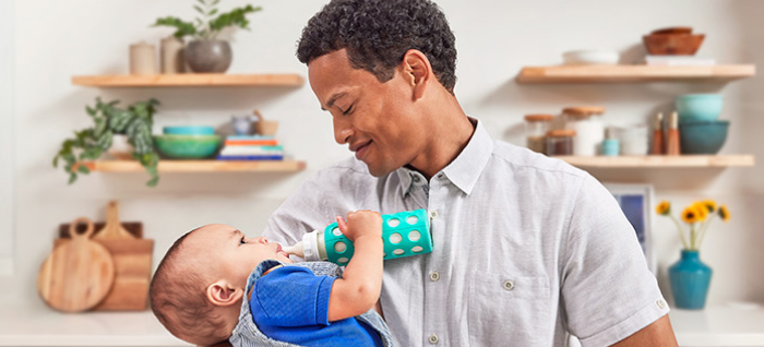 Man standing in kitchen and holding his baby boy, who is dressed in blue and drinking a bottle of EleCare.